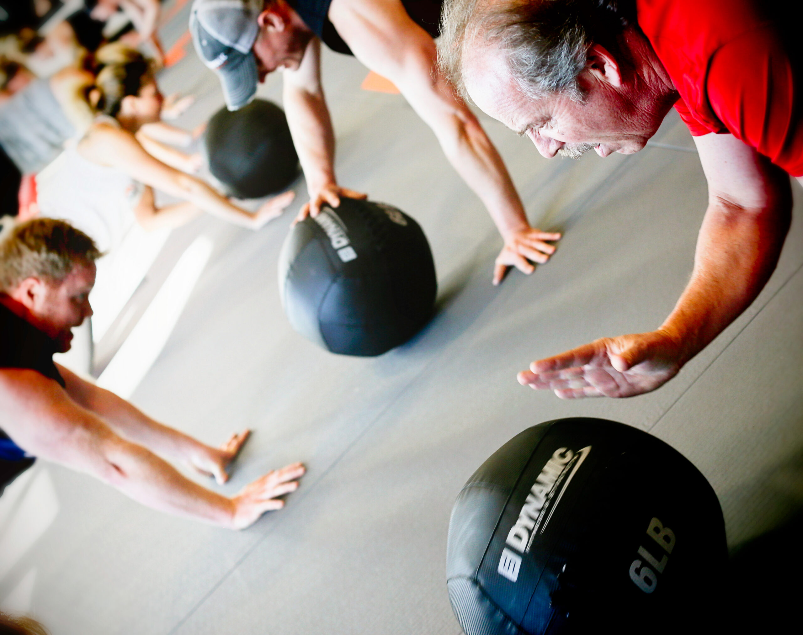 Two smiling Fitaholic Fitness members posing together after a workout, with gym equipment and other members in the background at a group fitness gym.